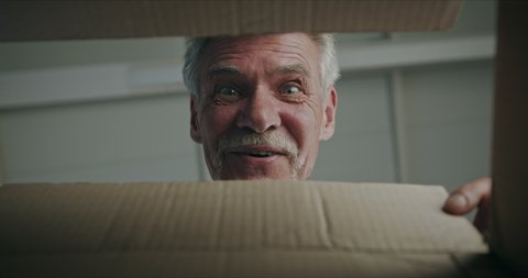 Joyful surprised gray-haired senior man pensioner opening cardboard box with surprise gift present on birthday special occasion, looking happy pleased and amazed.