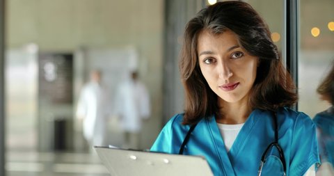 Portrait shot of Caucasian beautiful young woman doctor smiling at camera and holding folder. Close up of pretty happy female physician smile. Medic in uniform in clinic. Indoors.