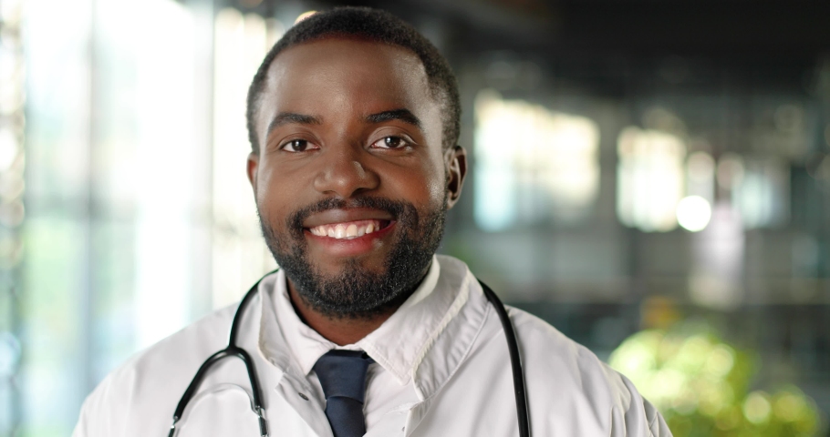Portrait of African American young man doctor with stethoscope smiling at camera cheerfully. Handsome happy male physician smile. Medic in white gown in clinic. Indoor. | Shutterstock HD Video #1059250142