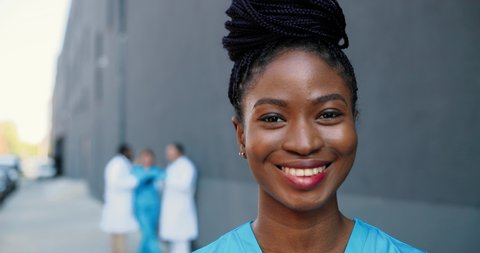 Portrait of African American young beautiful woman doctor looking at camera and smiling cheerfully. Pretty female physician smile. Multi ethnic doctors on background. Medic in white gown. Close up.