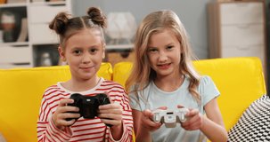 Portrait of the two cheerful and cute Caucasian small sisters sitting on the yellow couch and playing videogames with joysticks in hands in cozy living room.