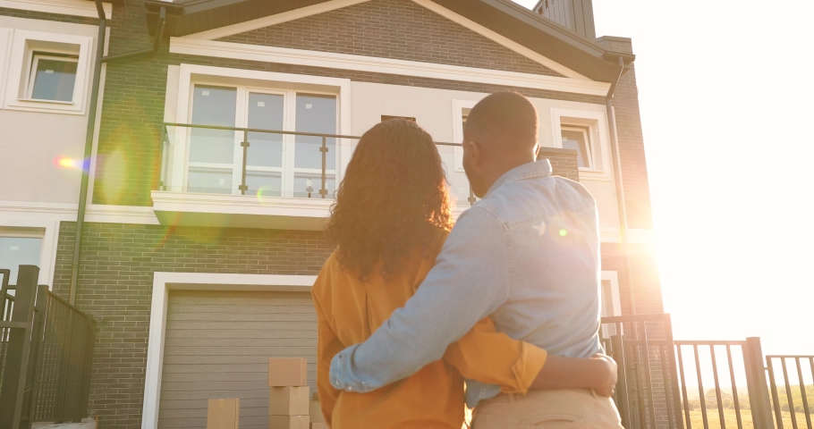 Rear on African American young just-married couple standing at yard of house, hugging and observing second floor. Back view on man and woman in hugs moving in new home. | Shutterstock HD Video #1059250358