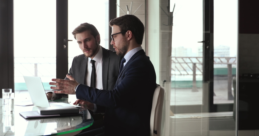 Two focused businessmen in formal wear discussing project presentation on computer, shaking hands after making agreement. Financial advisor handshaking with client after presenting profitable offer. Royalty-Free Stock Footage #1059252539
