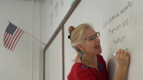 Pretty blond woman teaching spanish in a school on a whiteboard with US flag hanging.