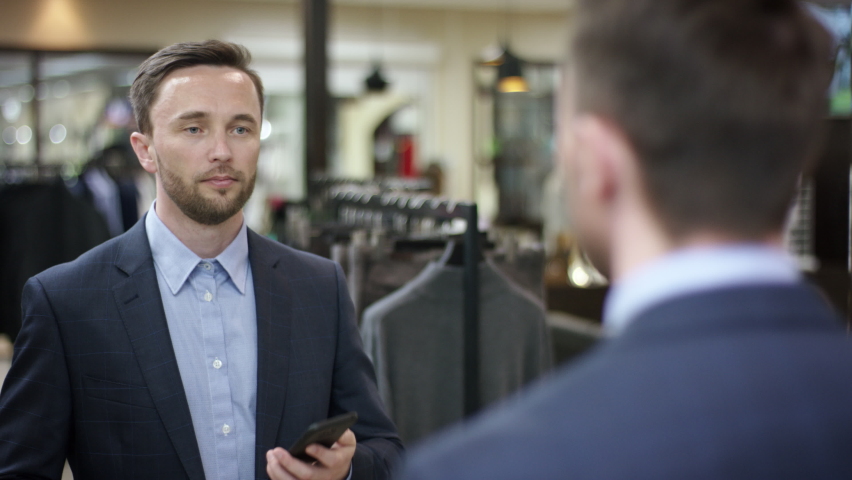 A close up shot over the shoulder of a middle-aged business man standing in front of the mirror trying on a new jacket and talking with his partner on the phone