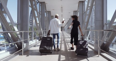 Back view of African young woman and American black man walk together talking, mixed race couple carry suitcases in airport terminal or train station, travel vacation together friends or marrieds