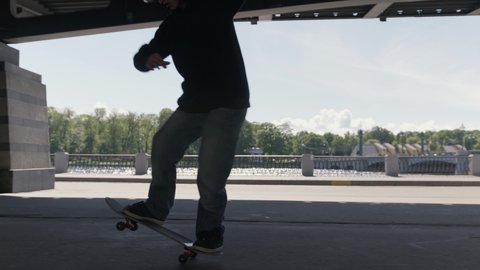  Teenager rides on back wheels of skateboard performing balance tricks on site of skate Park under an iron bridge. Professional skateboarder guy in dark clothing performs an trick on skateboard