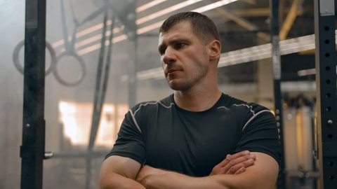 Portrait of a strong athletic man in a closed pose looking at the camera. Interior of a modern sports hall at sunset, professional studio lighting, serious epic decisive look.