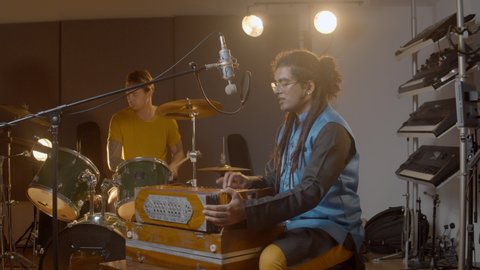 Music recording. Indian man playing harmonium in the recording studio with caucasian man on drums. Hindi male person in national clothes plays meditation music.