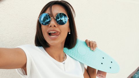 Young beautiful sexy smiling hipster woman in sunglasses.Trendy girl in summer T-shirt and shorts.Positive female with blue penny skateboard posing in the street near white wall. Taking selfie photos