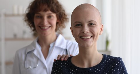 Middle-aged woman oncology nurse practitioner standing behind cancer patient, smile look at camera, head shot portrait. Doctor supporting ill female put hand on shoulder as symbol of help and