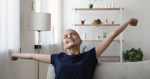Young bald woman stretch arms resting on couch alone. Hairless female put hands behind head enjoy free time, breath fresh air feels alive. No stress, remission day of illness. Cancer patient concept.