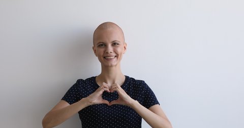 Bald young attractive female portrait on gray studio background feels healthy looks at camera showing heart symbol with hands. Cancer patient in love with life, healed woman express gratitude concept
