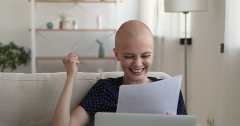 Young bald after chemotherapy woman cancer patient female sitting on couch at home holding paper sheets feels happy woman reading test about health results. Remission oncology disease recovery concept