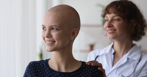 Bald 20s female look at window dream about healing. Middle aged stand near her put hand on shoulder gives care show empathy, provide psychological support to patient during cancer rehabilitation