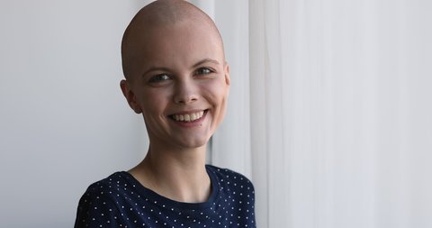 Bald cancer patient standing near window looking outside thinking about therapy. Woman feel optimistic smile look at camera. Hair loss after chemotherapy, oncology disease recovery, rehab concept
