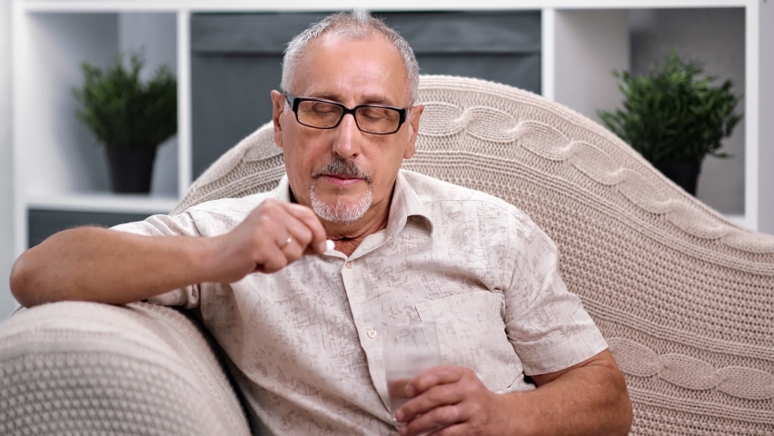 Unwell mature 70s grandfather take pill drinking water having health problem or chronic illness. Unhappy sick senior man holding painkiller to relieve pain sitting on couch. Medium shot on RED camera | Shutterstock HD Video #1059275636