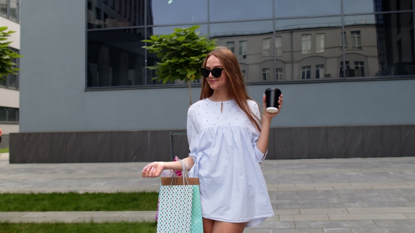 Girl staying near shopping mall with shopping bags and drinking coffee or tea. Looking satisfied and happy with purchase, enjoying discounts, low prices on Black Friday sales holiday. 6k downscale