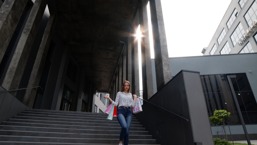 Cheerful pretty smiling girl walking from centre mall with colorful shopping bags, looking satisfied and happy with purchase, enjoying discounts, low prices on Black Friday sales holiday. 6k downscale