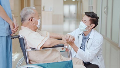 Nurse push Asian senior adult patient wheelchair in hospital hallway, young male doctor crouch down holding hand and talk to the patient with care. Medical healthcare job, or hospital business concept