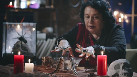 Portrait of old gypsy witch casting spell using two serpents, burning candles and bowl of water. Fortune teller performing ritual to foresee future
