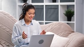 Happy young woman in headphones talking online video call use laptop sitting on couch at home. Smiling female gesturing working remotely enjoying friendly conversation. Medium shot on RED camera