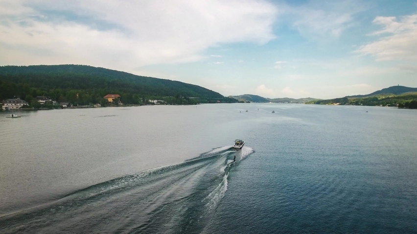 Aerial drone tracking shot of a speed boat and a man waterskiing doing water sports in Velden Am Worthersee lake, Austria on a beautiful sunny day mountain view Royalty-Free Stock Footage #1059281318