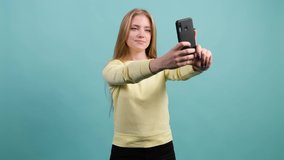 Funny young blonde haired woman making selfie. Smiling girl holding smartphone, making faces on camera, isolated on turquoise background.
