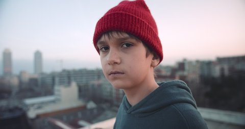 Handsome trendy young boy on rooftop at sunset wearing hoody and beanie red hat looking confident in urban city background.