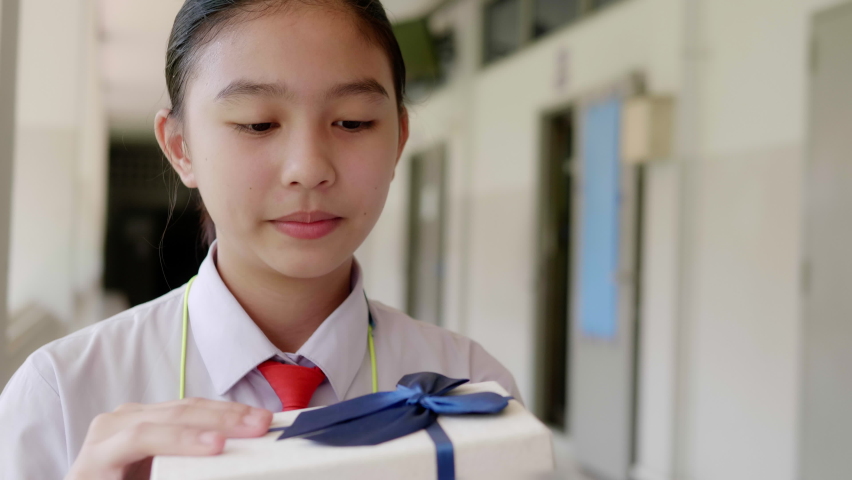 A smiling Asian teenage high school girl in a white school uniform opens a gift box and is surprised, excited and delighted to see the gift.