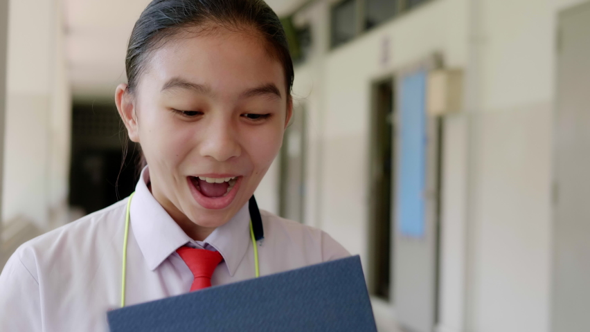 A smiling Asian teenage high school girl in a white school uniform opens a gift box and is surprised, excited and delighted to see the gift.