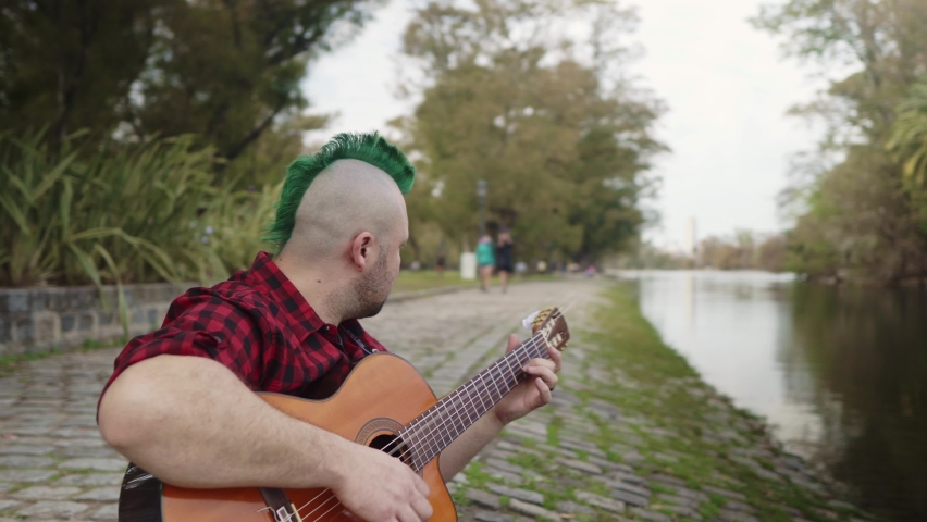 Dolly left shot of a caucasian alternative man playing guitar beside lake at park