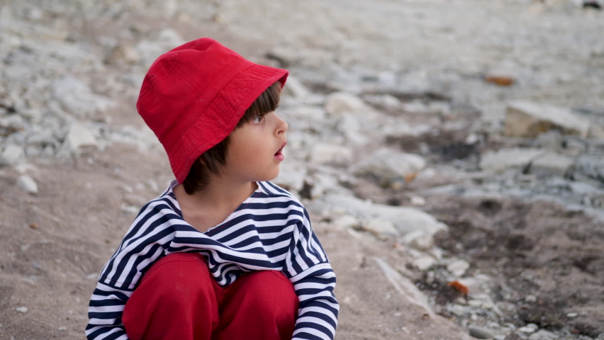 mom adjusts the hair of a boy child sitting on a stone beach in a red cap and vest