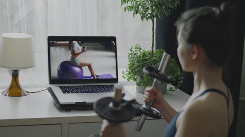 Asian woman doing sports and fitness training at home during covid quarantine. Young fit sporty brunette girl pumping muscles with dumbbells watching tutorial. Healthy lifestyle in pandemic time