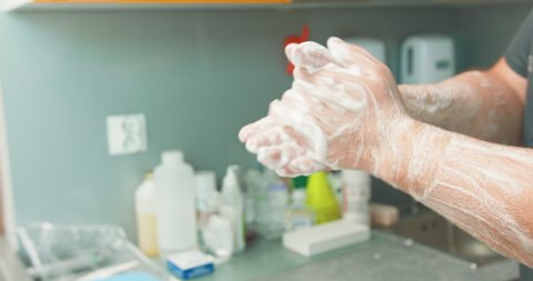 Close up hands of male veterinarian, in foam, then he turns to the tap, while he washes hands before an operation. At the background - blurred wall and a table with medical polymeric containers.