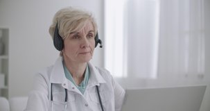 aged female family doctor is consulting online, talking with patient by video chat at laptop, working remotely