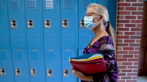 Side view of teacher walking down a hallway in an empty school wearing a medical face mask and holding books showing emptiness.