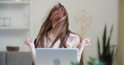 Active young girl freelancer student sits at desk in front of laptop, dances, moves waves her arms and shakes head and hair, feels joy enjoyment of freedom, relaxes on break, funny frenzied movements