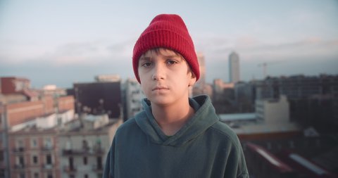 Portrait young boy on rooftop at sunset wearing hoody and beanie red hat looking confident in urban city background