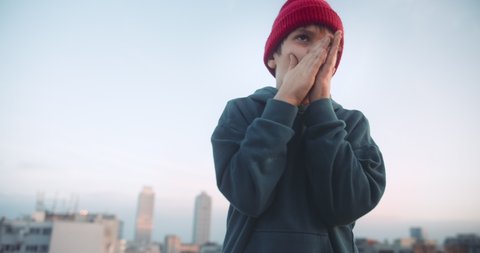 Handsome young boy dancing and have fun on rooftop at sunset. Wearing hoody and beanie red hat looking confident in urban city background