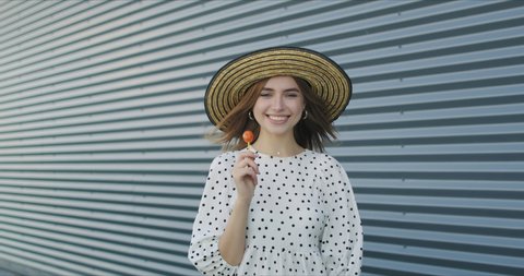 Fashion portrait sweet young woman having fun with lollipop over wall background. Pretty girl in dress and hat. 4k raw video footage