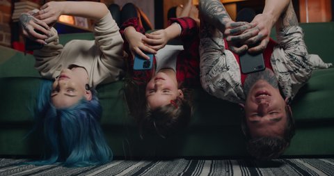 Cheerful family lying on sofa upside down and using smartphones. Modern mother, father and their kid chatting in social networks while spending time. People with tattoos and piercing.