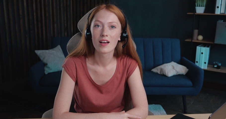 Female Ginger with Headset is talking to the Camera, recording Vlog, Blog. A  red-haired Woman is holding Webinar, Video-Conference, Video-Call. Distance Job, Studying, Education. Student Indoor. Royalty-Free Stock Footage #1059311276
