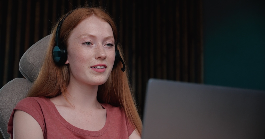 Smiling Woman with Headset using a Laptop, talking. Young Ginger Woman is working as a Customer support service Operator at the Home office in Headphones with a Microphone. Distance Job, Lifestyle. Royalty-Free Stock Footage #1059311285
