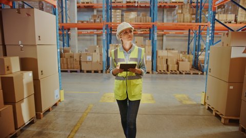 Professional Female Worker Wearing Hard Hat Uses Digital Tablet Checks Inventory Walks in the Retail Warehouse full of Shelves with Goods. Working in Logistics, Distribution Center. Following Shot