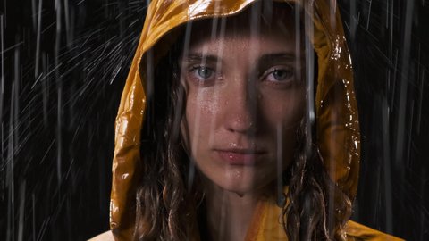Close-up footage of beautiful woman with green eyes who is standing in the pouring rain, looking at the camera and wearing yellow raincoat hood. Nice lady looks straight into the camera, rack focus.