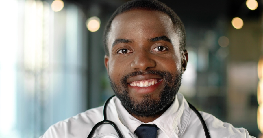 Portrait of African American young man doctor with stethoscope looking at camera and smiling cheerfully. Handsome happy male physician smile. Medic in white gown in clinic. Indoor. | Shutterstock HD Video #1059319286