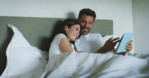 Authentic shot of young happy smiling just married couple in love is enjoying time together while using tablet for family entertainment on a bed in a bedroom just woke up in the morning in a sunny day