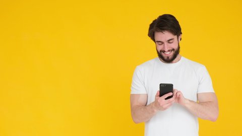 Handsome unshaven bearded young guy 20s wearing white t-shirt isolated over pastel blue background in studio. People sincere emotions lifestyle concept. Looking surprised wow, using mobile cell phone.