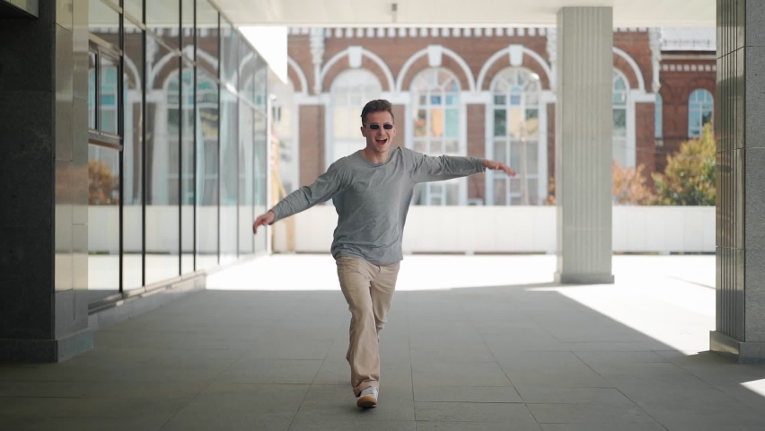 Handsome happy man in sunglasses is actively running, smiling and dancing with joy and happiness along the city street. Scene Shot in an Urban Park Next to Business Center. Slow motion.